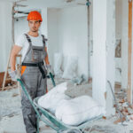 A young Caucasian male construction worker is using a construction trolley to take out bags of renovation residue.